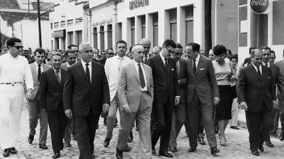 Fotografia antiga em preto e branco mostra o líder político doutor Álvaro Berardinelli de terno claro caminhando com comitiva sobre paralelepípedos em Mendes.