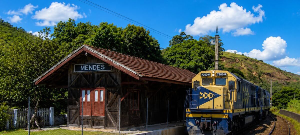 Fotografia da histórica Estação Ferroviária Neri Ferreira feita de madeira escura com placa identificadora escrita Mendes ao lado de uma moderna locomotiva amarela e azul da empresa MRS Logística estacionada nos trilhos sob céu azul com nuvens.