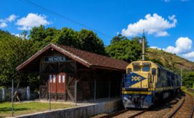 Fotografia da histórica Estação Ferroviária Neri Ferreira feita de madeira escura com placa identificadora escrita Mendes ao lado de uma moderna locomotiva amarela e azul da empresa MRS Logística estacionada nos trilhos sob céu azul com nuvens.