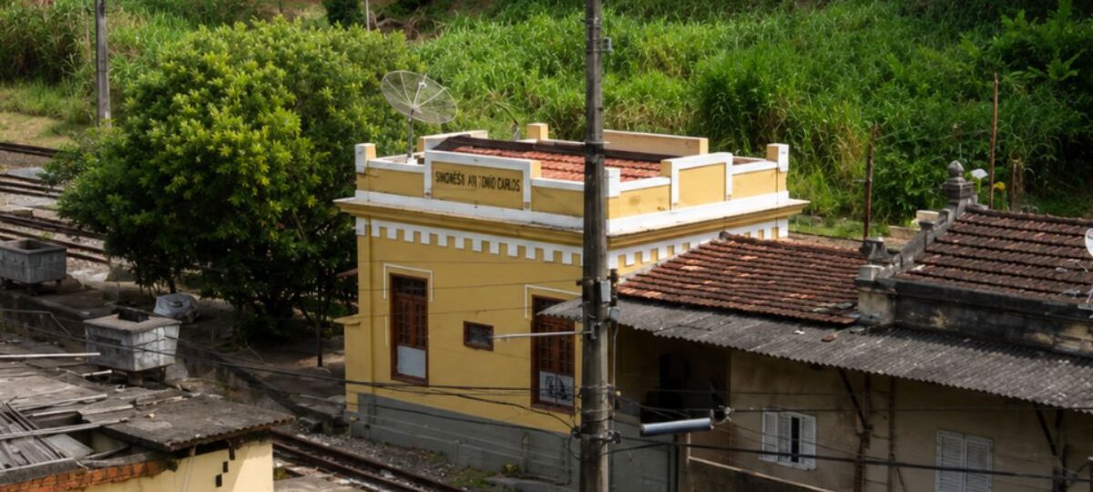 Fachada da histórica estação de Humberto Antunes com paredes amarelas cercada por vegetação e trilhos de trem no município de Mendes.