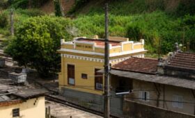 Fachada da histórica estação de Humberto Antunes com paredes amarelas cercada por vegetação e trilhos de trem no município de Mendes.