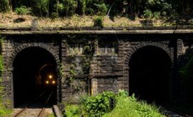 Fotografia com vista frontal das duas entradas de pedra escura dos Túneis 12 e 12 Bis em Mendes, com trilhos ferroviários e os faróis acesos de um trem surgindo no interior da galeria esquerda, cercada por vegetação verde na Serra do Mar.