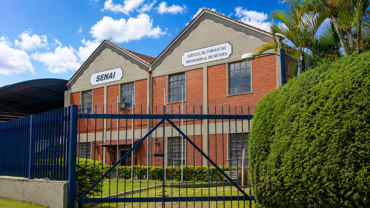 Fachada externa do prédio do SENAI em Mendes com paredes de tijolos aparentes vermelhos, portão de metal azul na frente, jardins verdes e céu azul com nuvens.