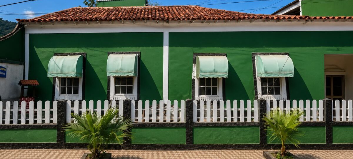 Fotografia da fachada frontal da Casa do Barão de Santa Cruz em Mendes, Rio de Janeiro, exibindo paredes pintadas de verde com detalhes brancos, quatro janelas coloniais quadradas com toldos claros arqueados, telhado de telhas cerâmicas coloniais, cercada por uma mureta verde e preta com grade de madeira branca, além de duas pequenas palmeiras plantadas na calçada de piso decorado.