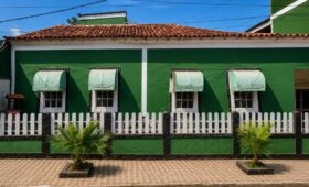 Fotografia da fachada frontal da Casa do Barão de Santa Cruz em Mendes, Rio de Janeiro, exibindo paredes pintadas de verde com detalhes brancos, quatro janelas coloniais quadradas com toldos claros arqueados, telhado de telhas cerâmicas coloniais, cercada por uma mureta verde e preta com grade de madeira branca, além de duas pequenas palmeiras plantadas na calçada de piso decorado.