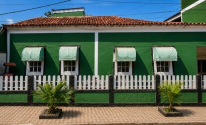 Fotografia da fachada frontal da Casa do Barão de Santa Cruz em Mendes, Rio de Janeiro, exibindo paredes pintadas de verde com detalhes brancos, quatro janelas coloniais quadradas com toldos claros arqueados, telhado de telhas cerâmicas coloniais, cercada por uma mureta verde e preta com grade de madeira branca, além de duas pequenas palmeiras plantadas na calçada de piso decorado.