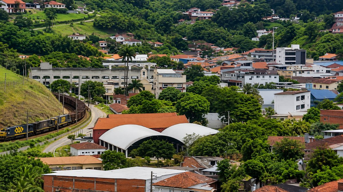 Vista aérea da cidade de Mendes no estado do Rio de Janeiro mostrando casas entre colinas verdes e um trem de carga circulando pelas ferrovias locais.