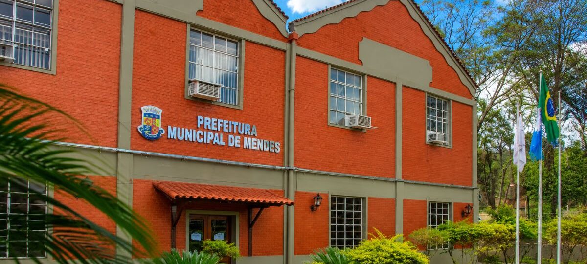 Fotografia colorida da fachada de um imponente prédio histórico de dois andares com paredes de tijolos vermelhos aparentes e detalhes estruturais em tom verde oliva, onde funciona atualmente a Prefeitura Municipal de Mendes no Rio de Janeiro. A fachada exibe grandes janelas quadradas de vidro com molduras brancas, aparelhos de ar condicionado instalados, o brasão oficial do município ao lado das letras em relevo azul e branco com a inscrição Prefeitura Municipal de Mendes, e um jardim gramado em frente com pequenos arbustos decorativos e três mastros com bandeiras hasteadas sob um céu azul ensolarado com nuvens brancas.