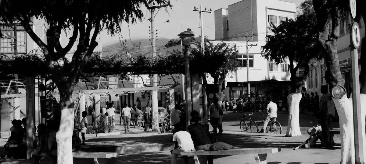 Fotografia antiga em preto e branco mostra a Rua Francisco Cabral em Mendes, com pessoas sentadas em bancos de uma praça arborizada e pedestres ao fundo circulando perto de prédios.