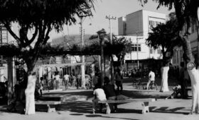 Fotografia antiga em preto e branco mostra a Rua Francisco Cabral em Mendes, com pessoas sentadas em bancos de uma praça arborizada e pedestres ao fundo circulando perto de prédios.