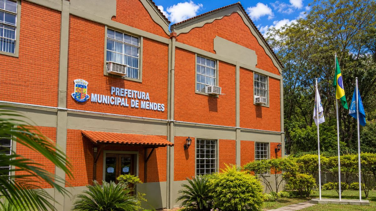 Fachada do prédio de tijolos vermelhos da Prefeitura Municipal de Mendes com letreiro oficial, brasão colorido, bandeiras hasteadas e jardins verdes ao redor sob céu azul.