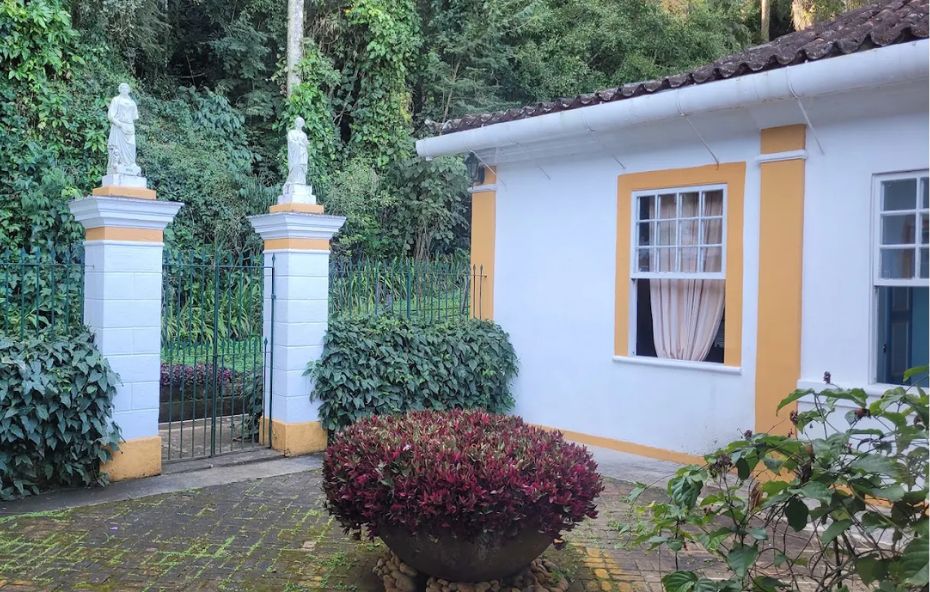 Fotografia da entrada de uma construção colonial branca com detalhes em amarelo ocre onde se destacam duas colunas brancas com estátuas clássicas no topo ladeando um portão de ferro verde e um grande vaso de plantas ornamentais em primeiro plano.