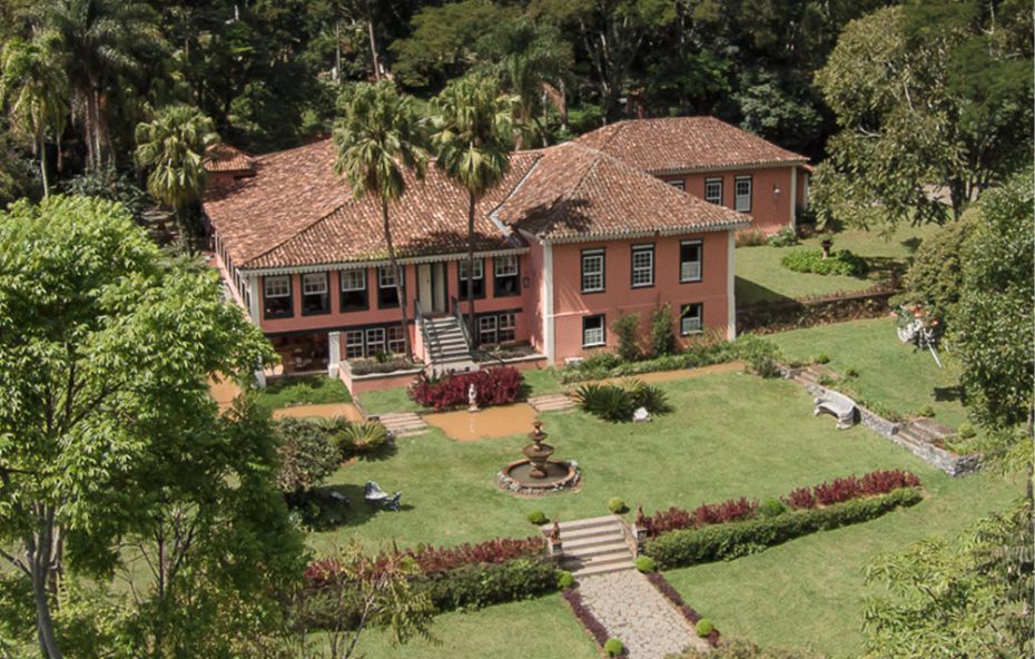Vista aérea de um imponente casarão histórico de cor rosada com detalhes em branco e telhado de barro cercado por jardins simétricos com palmeiras imperiais e uma fonte central em meio a uma densa vegetação de mata atlântica no Rio de Janeiro.