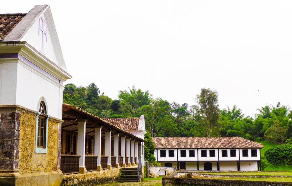 Fotografia da fachada lateral de um casarão colonial histórico com paredes brancas e detalhes em pedra amarela sob um telhado de barro antigo. A imagem mostra uma varanda extensa com colunas brancas e um pátio de pedra que leva a outra construção de dois andares ao fundo cercada por uma densa vegetação verde e árvores altas.