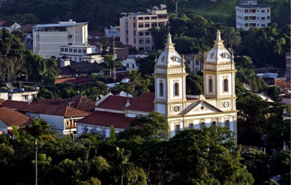 Fotografia aérea da Catedral de Nossa Senhora da Glória em Valença, Rio de Janeiro. A imponente igreja branca de estilo colonial, com suas duas torres, se destaca em meio à vegetação densa e às construções da cidade ao fundo. A luz do sol ilumina a fachada da igreja, ressaltando seus detalhes arquitetônicos.