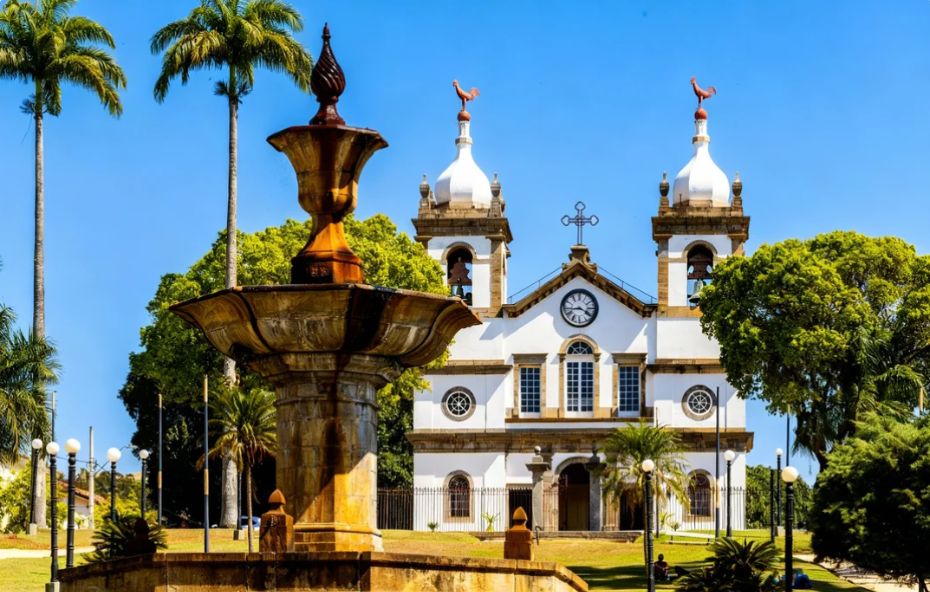 Fotografia da Igreja Matriz de Nossa Senhora da Conceição em Vassouras, Rio de Janeiro. A igreja branca de estilo colonial está ao fundo em um gramado. Em primeiro plano, um grande chafariz de pedra se destaca. O céu está azul e há palmeiras e outras árvores ao redor.