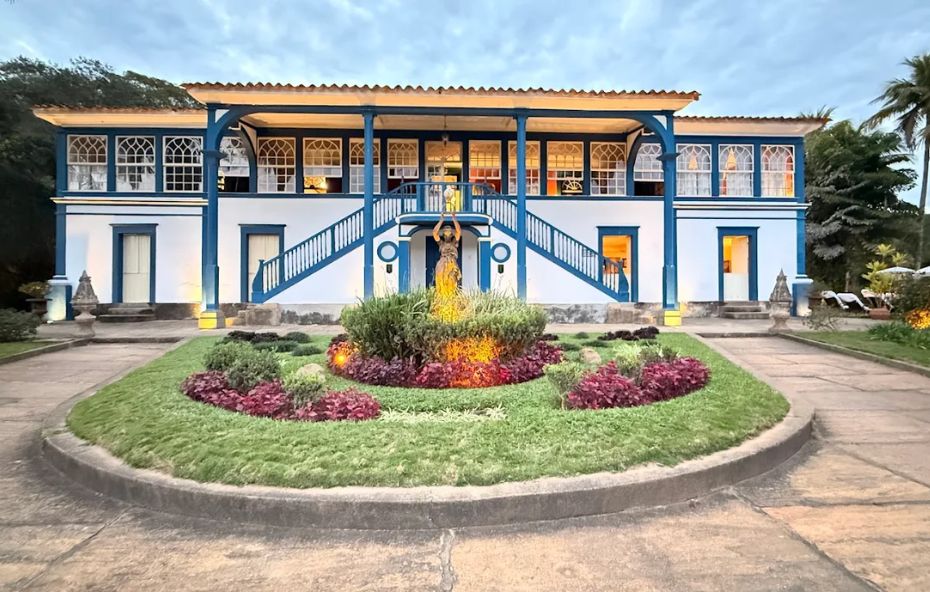Fotografia de uma imponente casa colonial de dois andares com fachadas brancas e detalhes em azul intenso destacando as janelas e a escadaria central enquanto um jardim meticulosamente cuidado com flores coloridas ocupa o primeiro plano sob um céu de fim de tarde.