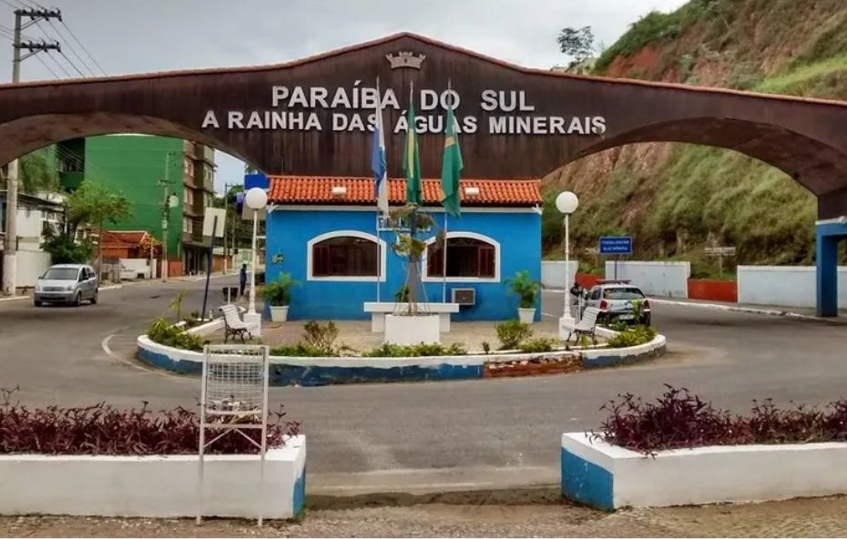 Fotografia do pórtico de entrada da cidade de Paraíba do Sul. Um grande arco marrom com os dizeres "Paraíba do Sul, A Rainha das Águas Minerais" se estende sobre uma rotatória e uma guarita azul.