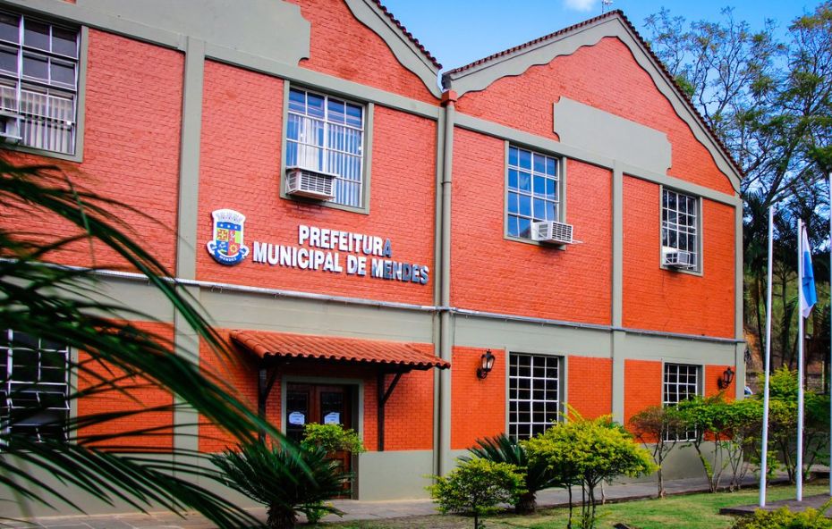 Fotografia da fachada da Prefeitura Municipal de Mendes, um prédio histórico de tijolos vermelhos que abrigou a antiga Cervejaria Teutônia. O brasão e o nome da cidade estão na parede. À direita, bandeiras hasteadas e, ao redor, árvores e vegetação.