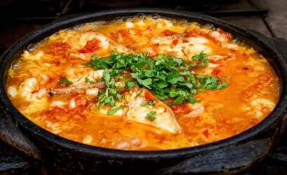 Fotografia em close de uma moqueca de peixe fervendo em uma panela de barro preta. O caldo é alaranjado e rústico, com pedaços visíveis de peixe branco, tomate e cebola. No centro, há uma porção generosa de coentro fresco picado sobre o ensopado.