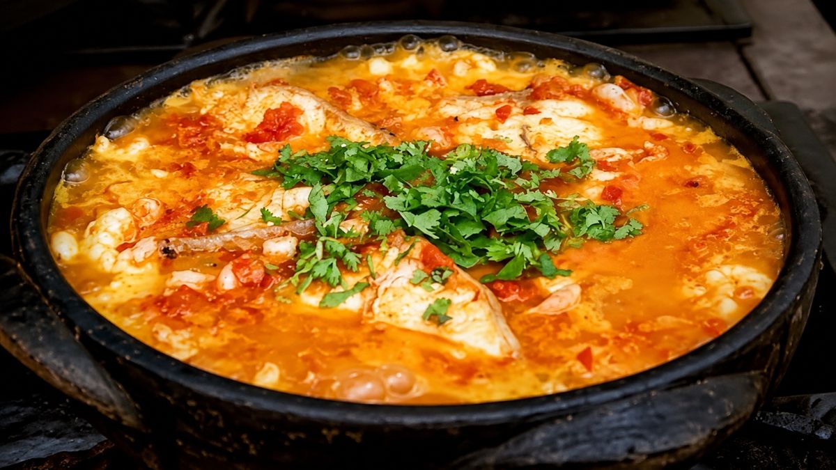 Fotografia em close de uma moqueca de peixe fervendo em uma panela de barro preta. O caldo é alaranjado e rústico, com pedaços visíveis de peixe branco, tomate e cebola. No centro, há uma porção generosa de coentro fresco picado sobre o ensopado.