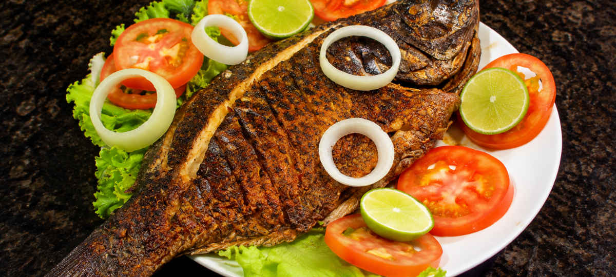 Peixe inteiro dourado e assado na brasa, servido em um prato branco sobre folhas de alface verde, decorado com rodelas vermelhas de tomate, anéis de cebola branca e fatias redondas de limão verde fresco.