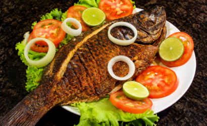 Peixe inteiro dourado e assado na brasa, servido em um prato branco sobre folhas de alface verde, decorado com rodelas vermelhas de tomate, anéis de cebola branca e fatias redondas de limão verde fresco.