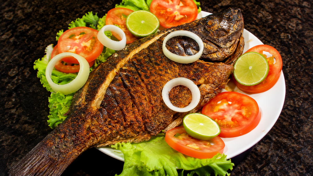 Peixe inteiro dourado e assado na brasa, servido em um prato branco sobre folhas de alface verde, decorado com rodelas vermelhas de tomate, anéis de cebola branca e fatias redondas de limão verde fresco.