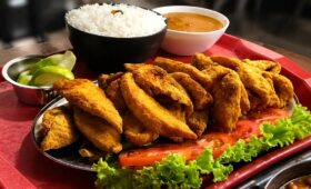 Uma travessa com filés de peixe frito dourado e crocante servida com acompanhamentos como arroz branco, pirão cremoso e uma salada de alface e tomate frescos, tudo em uma bandeja vermelha sobre uma mesa de madeira.
