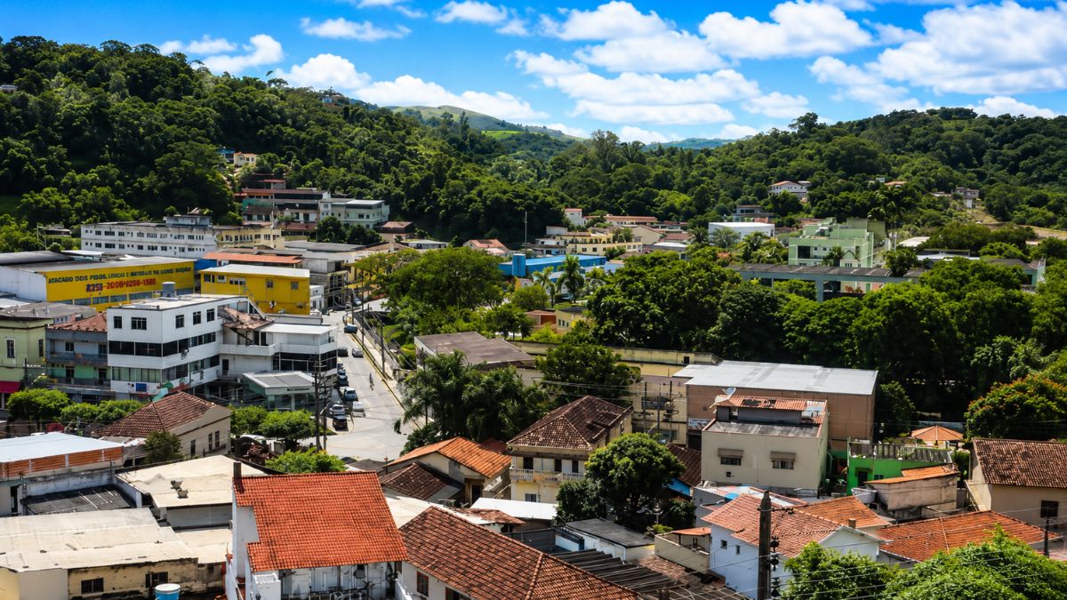 Fotografia panorâmica de cima mostrando o centro da cidade de Mendes no Rio de Janeiro com casas de telhado colonial, prédios comerciais de poucos andares, ruas com carros e colinas cobertas por mata densa sob céu azul com nuvens brancas.