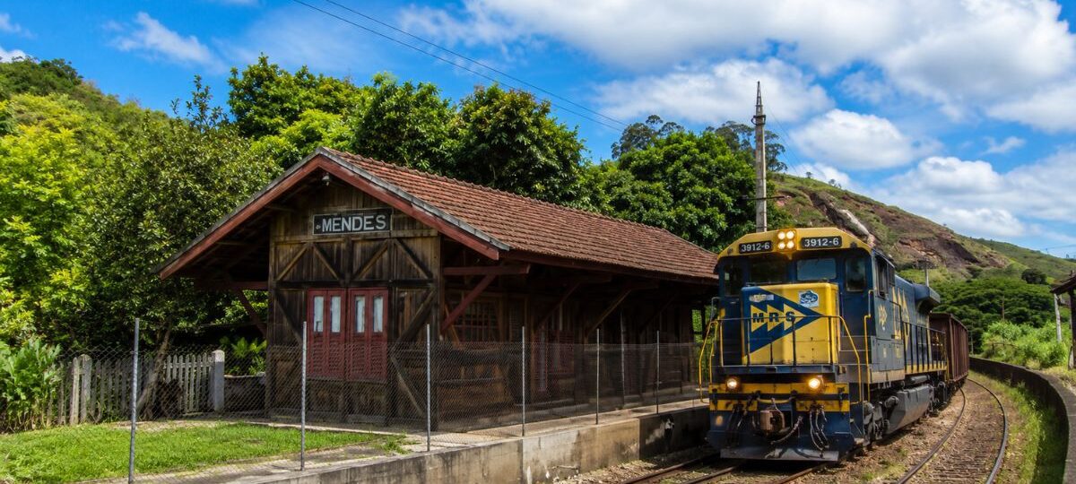 Estação Ferroviária Neri Ferreira em Mendes, Rio de Janeiro, com prédio de madeira histórico e locomotiva azul da MRS passando nos trilhos.