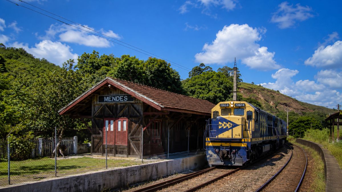 Estação Ferroviária Neri Ferreira em Mendes RJ com estrutura antiga de madeira rústica e trem moderno azul e amarelo sobre os trilhos.