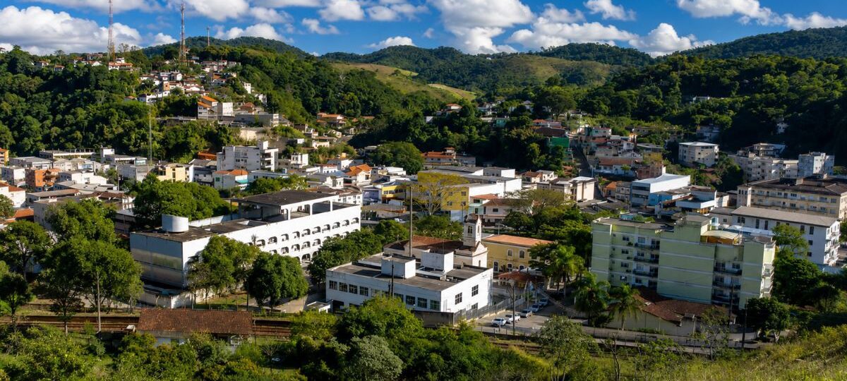 Vista aérea em plano médio da cidade de Mendes, no Rio de Janeiro, exibindo prédios brancos de arquitetura predominantemente moderna e a Igreja Matriz, aninhados em um vale cercado por colinas densamente cobertas por vegetação nativa da Mata Atlântica e morros mais distantes com algumas habitações. No topo de uma das colinas à esquerda, torres de transmissão são visíveis contra um céu azul brilhante com nuvens brancas e macias.