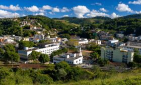 Vista aérea em plano médio da cidade de Mendes, no Rio de Janeiro, exibindo prédios brancos de arquitetura predominantemente moderna e a Igreja Matriz, aninhados em um vale cercado por colinas densamente cobertas por vegetação nativa da Mata Atlântica e morros mais distantes com algumas habitações. No topo de uma das colinas à esquerda, torres de transmissão são visíveis contra um céu azul brilhante com nuvens brancas e macias.