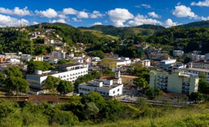 Vista aérea em plano médio da cidade de Mendes, no Rio de Janeiro, exibindo prédios brancos de arquitetura predominantemente moderna e a Igreja Matriz, aninhados em um vale cercado por colinas densamente cobertas por vegetação nativa da Mata Atlântica e morros mais distantes com algumas habitações. No topo de uma das colinas à esquerda, torres de transmissão são visíveis contra um céu azul brilhante com nuvens brancas e macias.