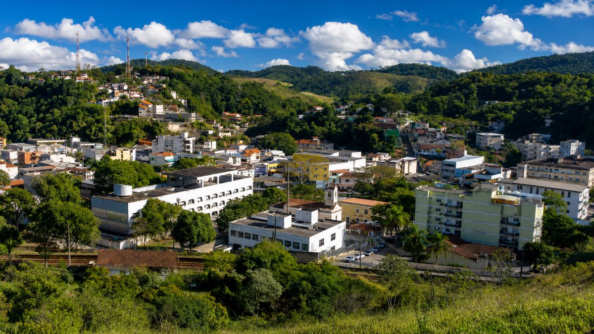Vista aérea em plano médio da cidade de Mendes, no Rio de Janeiro, exibindo prédios brancos de arquitetura predominantemente moderna e a Igreja Matriz, aninhados em um vale cercado por colinas densamente cobertas por vegetação nativa da Mata Atlântica e morros mais distantes com algumas habitações. No topo de uma das colinas à esquerda, torres de transmissão são visíveis contra um céu azul brilhante com nuvens brancas e macias.