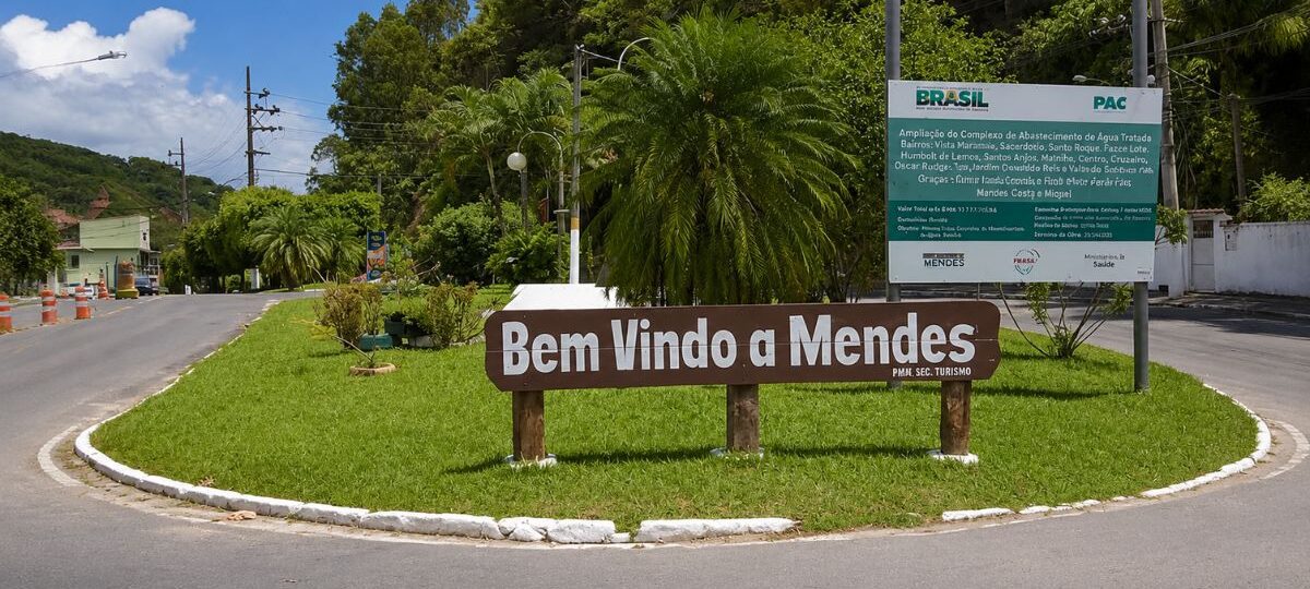 Placa de madeira marrom com a inscrição Bem Vindo a Mendes fixada em um canteiro gramado redondo na entrada do município fluminense.