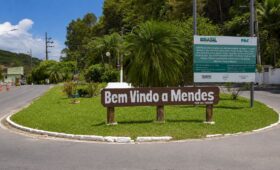 Placa de madeira marrom com a inscrição Bem Vindo a Mendes fixada em um canteiro gramado redondo na entrada do município fluminense.