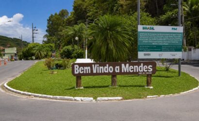 Placa de madeira marrom com a inscrição Bem Vindo a Mendes fixada em um canteiro gramado redondo na entrada do município fluminense.