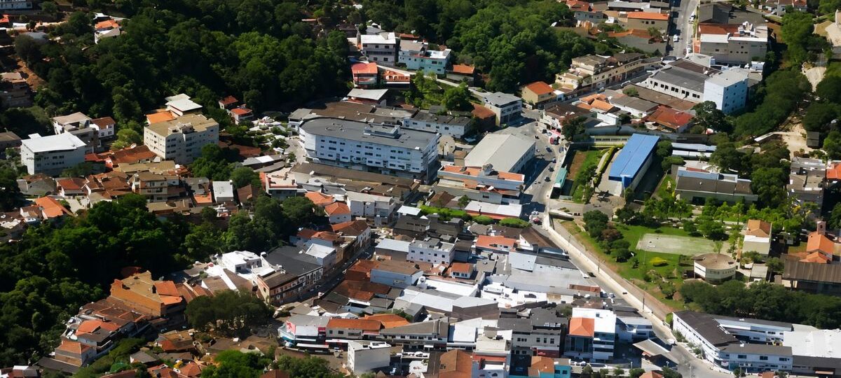 Fotografia aérea mostrando o centro urbano de Mendes com diversas casas, edifícios comerciais, ruas sinuosas e densa vegetação de Mata Atlântica ao fundo.