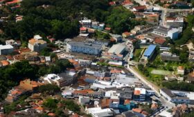 Fotografia aérea mostrando o centro urbano de Mendes com diversas casas, edifícios comerciais, ruas sinuosas e densa vegetação de Mata Atlântica ao fundo.