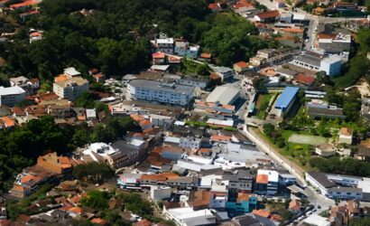 Fotografia aérea mostrando o centro urbano de Mendes com diversas casas, edifícios comerciais, ruas sinuosas e densa vegetação de Mata Atlântica ao fundo.