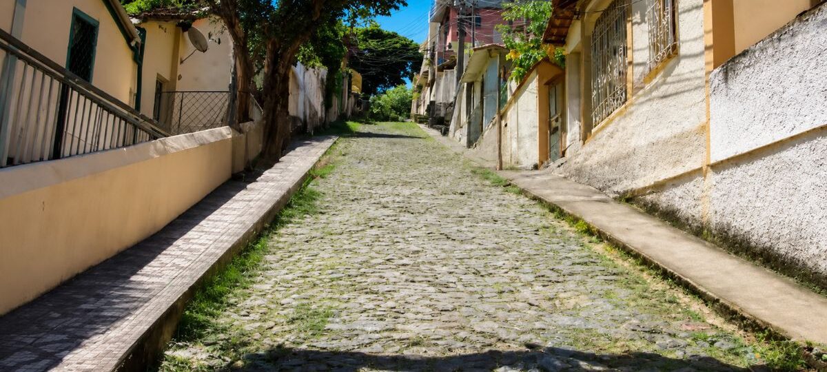 Fotografia da Ladeira João Vieira em Mendes com calçamento antigo de pedras irregulares estilo pé de moleque ladeada por residências e árvores sob céu azul límpido.