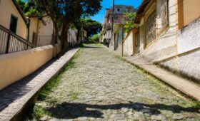 Fotografia da Ladeira João Vieira em Mendes com calçamento antigo de pedras irregulares estilo pé de moleque ladeada por residências e árvores sob céu azul límpido.