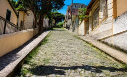 Fotografia da Ladeira João Vieira em Mendes com calçamento antigo de pedras irregulares estilo pé de moleque ladeada por residências e árvores sob céu azul límpido.