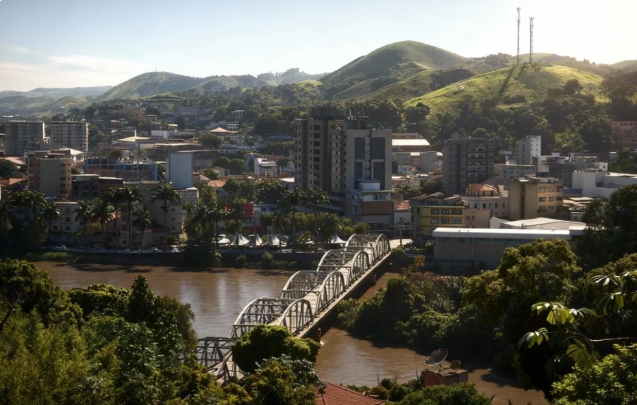 Vista panorâmica da cidade de Barra do Piraí, com a Ponte dos Arcos em destaque sobre as águas do Rio Paraíba do Sul. Ao fundo, prédios, casas e morros verdes compõem a paisagem sob um céu claro.