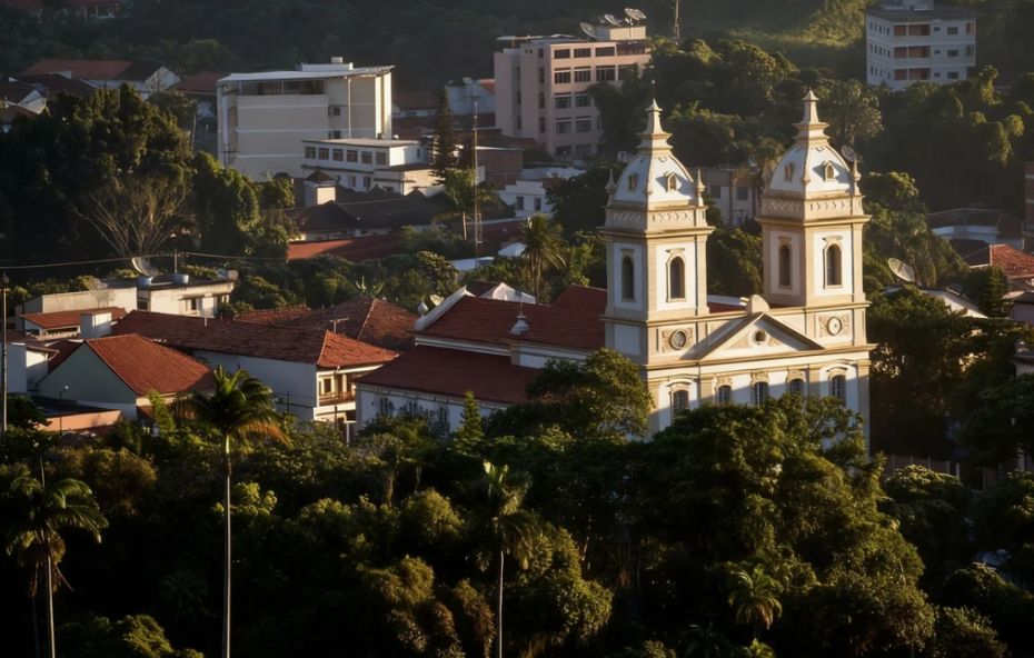 Vista da Catedral de Nossa Senhora da Glória em Valença, com suas duas torres brancas se destacando entre a vegetação verde e as casas da cidade ao redor, sob uma luz dourada.