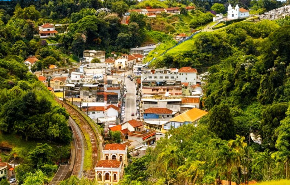 Vista aérea da cidade de Engenheiro Paulo de Frontin, aninhada em um vale com vegetação densa. É possível ver a rua principal, os trilhos de trem e uma igreja branca no alto de um morro.
