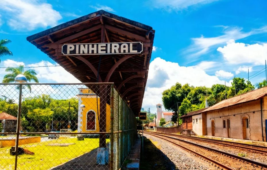Foto da plataforma da estação de trem de Pinheiral. A placa com o nome "PINHEIRAL" está em destaque sob o telhado de madeira. Os trilhos correm ao lado da plataforma, e ao fundo, há construções antigas e árvores.