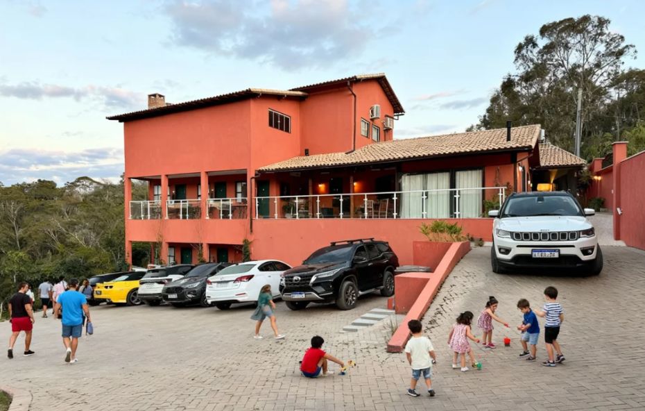 Edifício de dois andares com paredes em tom de terracota e telhado colonial onde carros estão estacionados e crianças brincam no pátio de pavimentação intertravada sob um céu de fim de tarde.